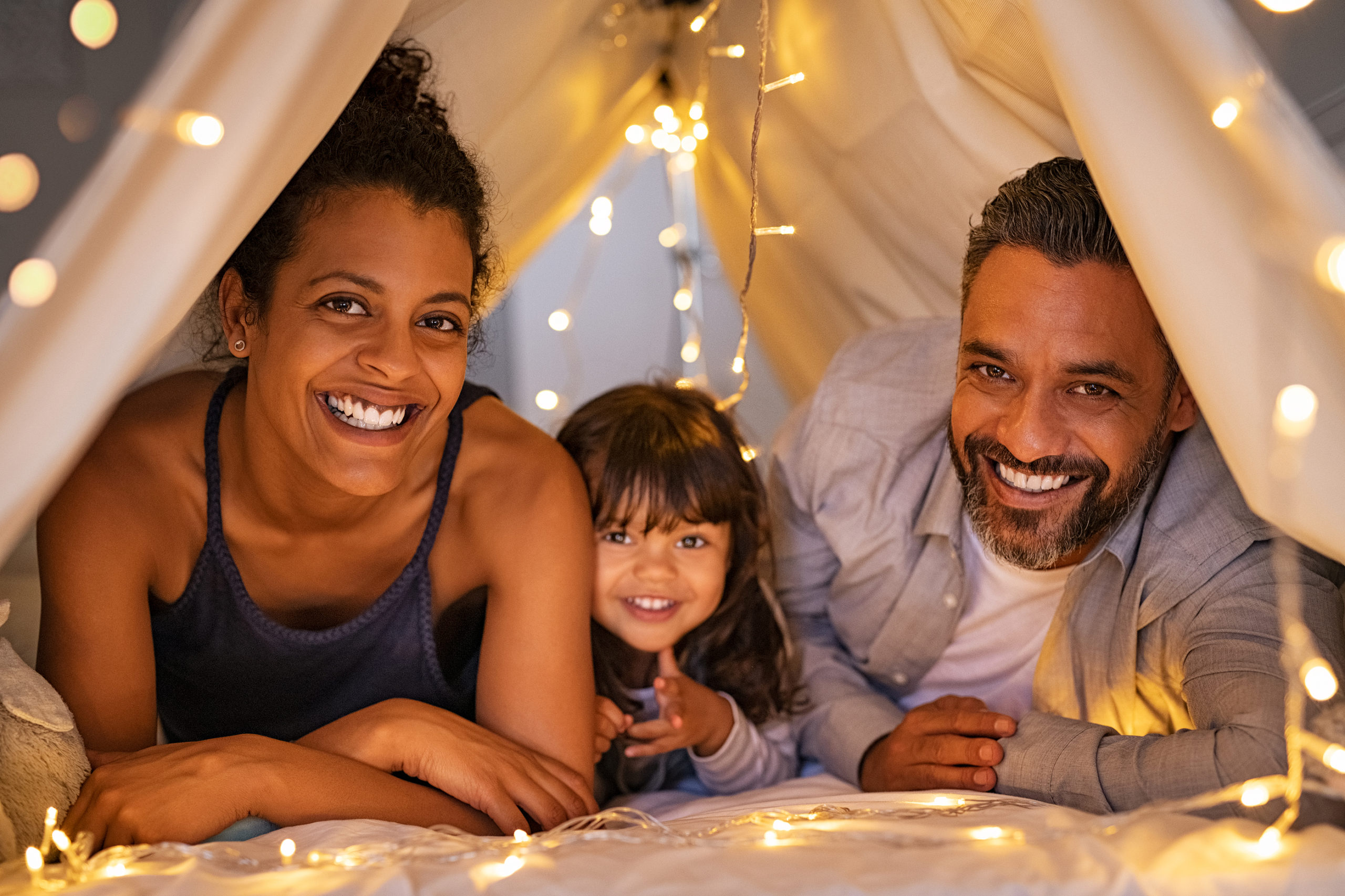 a happy mom and dad lay smiling with their daughter in a tent with fairy lights.