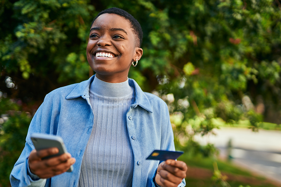 A young African American woman smiles outside while holding her smartphone and credit card